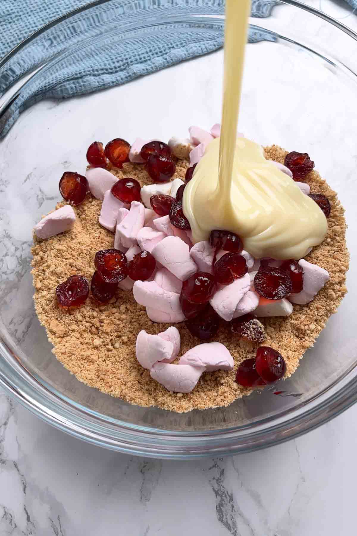 pouring condensed milk into a bowl of digestive biscuit crumbs, marshmallows and glace cherries
